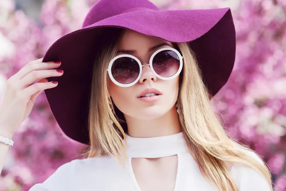 Stylish woman in purple hat and sunglasses posing outdoors with flowers.