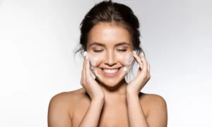 Smiling woman washing her face with foaming cleanser, eyes closed.