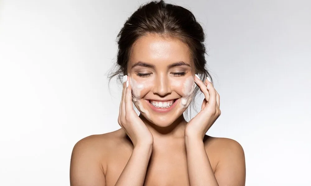 Smiling woman washing her face with foaming cleanser, eyes closed.