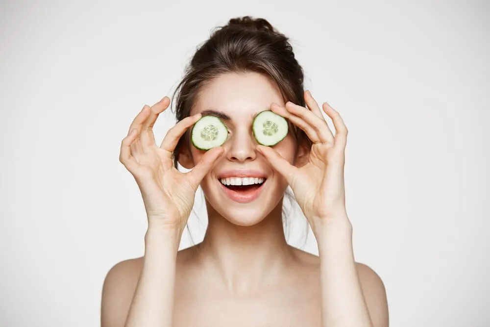 Smiling woman holding cucumber slices playfully over her eyes.