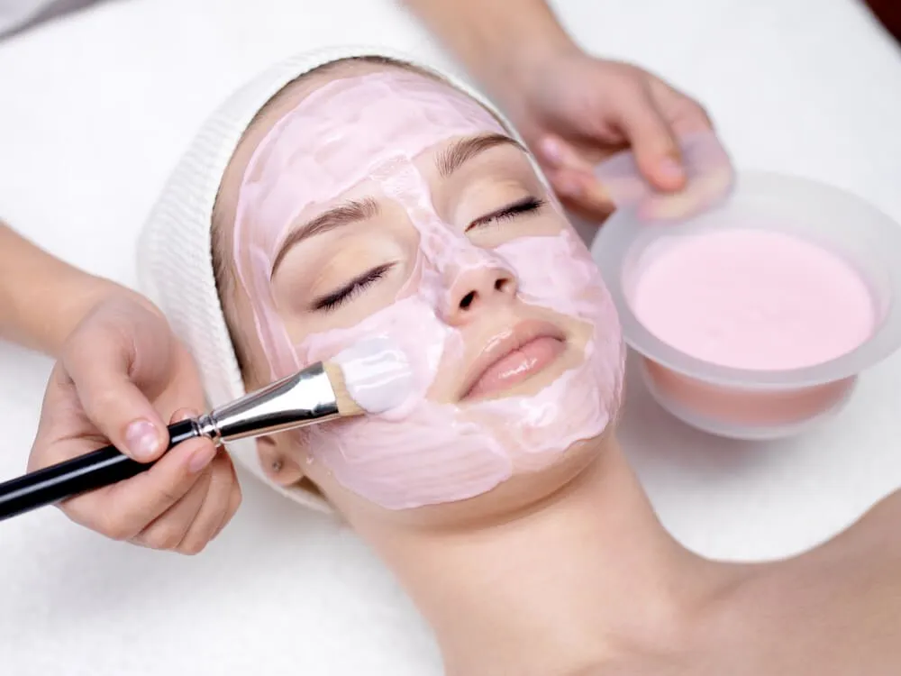 Person receiving spa treatment with pink facial mask applied by brush.