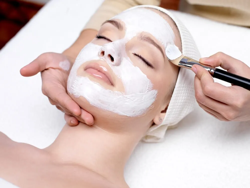 Woman lying down with white facial mask being applied by brush.